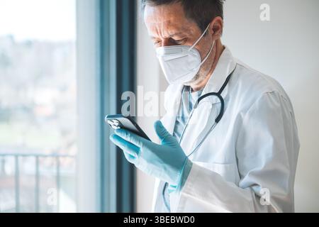 Stanco medico maschio esausto indossare maschera di protezione utilizzando smartphone in ospedale clinica ufficio, messa a fuoco selettiva Foto Stock