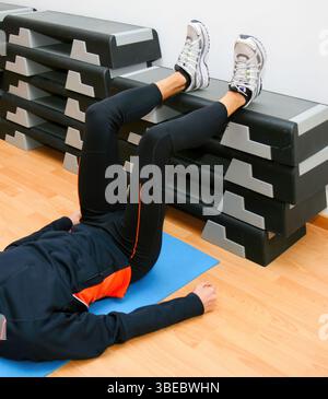 L'atleta giace su un tappetino da palestra, eseguendo un esercizio di sollevamento delle gambe con i piedi appoggiati su uno stepper, dimostrando forza e flessibilità in una routine di fitness Foto Stock