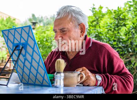 Vecchio con maglione rosso e capelli grigi che si radono seduti fuori nel suo giardino Foto Stock