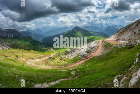 Tortuosa strada di montagna in una vibrante valle verde, vette alpine sotto un cielo spettacolare pieno di luci e ombre contrastanti in estate. Panora Foto Stock
