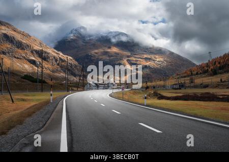 Strada asfaltata vuota che si snoda tra le montagne alpine tra nuvole al tramonto in autunno. Paesaggio con maestose Alpi, aranci ed erba, clou suggestivo Foto Stock