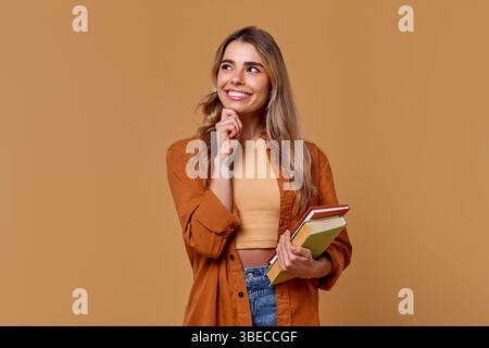 Una giovane donna allegra con lunghi fasci di capelli luminosi, mentre tiene una pila di libri colorati su uno sfondo caldo, che mostra il suo entusiasmo e. Foto Stock
