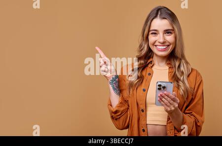 Una donna allegra con lunghi capelli irradia un sorriso luminoso, che punta felicemente su un lato mentre regge un elegante smartphone contro un retrogroun caldo e colorato Foto Stock