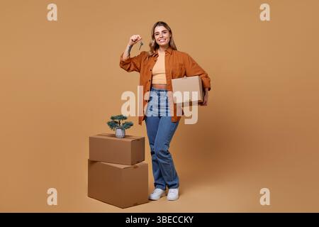 Una giovane donna allegra con una scatola e una chiave è circondata da scatole mobili e una pianta vivace, su un caldo sfondo beige, che incarna temi di vita Foto Stock