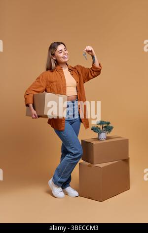 Una giovane donna allegra con una scatola e una chiave è circondata da scatole mobili e una pianta vivace, su un caldo sfondo beige, che incarna temi di vita Foto Stock