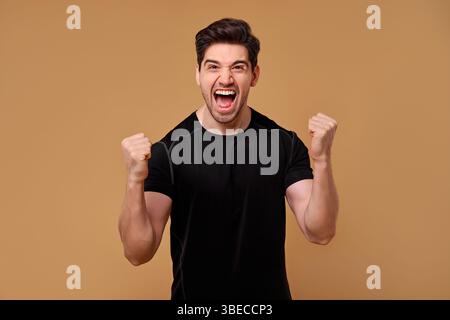 Un giovane con un sorriso ampio alza con gioia i pugni in alto per festeggiare, assaporando appieno la sua vittoria personale in un contesto neutrale che si Foto Stock