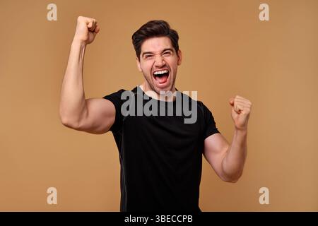 Un giovane con un sorriso ampio alza con gioia i pugni in alto per festeggiare, assaporando appieno la sua vittoria personale in un contesto neutrale che si Foto Stock