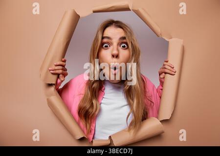 Una giovane donna dai capelli lunghi sbircia giocosamente attraverso la carta strappata, la sua espressione un delizioso mix di stupore e gioia, incarnando la curiosità giovanile Foto Stock
