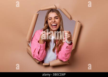 Una giovane donna allegra e vivace esplode energicamente attraverso un colorato sfondo di carta, che mostra il suo atteggiamento giocoso e la sua personalità dinamica, Foto Stock