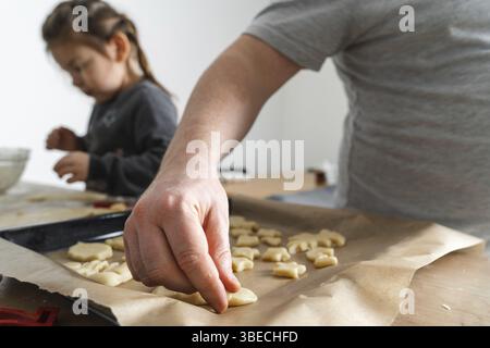 Vista ritagliata dell'uomo irriconoscibile e della figlia che fanno dolci con taglierine per biscotti sul pannello di lavorazione. Padre che mette i biscotti sulla teglia da forno Foto Stock