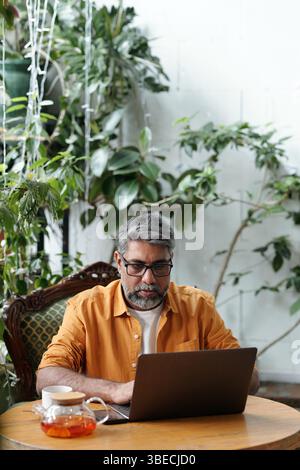 Uomo anziano con la barba che si aggancia al laptop mentre siede su una sedia circondato da lussureggianti piante interne, indossa occhiali e camicia arancione, che sembra contemplativo Foto Stock