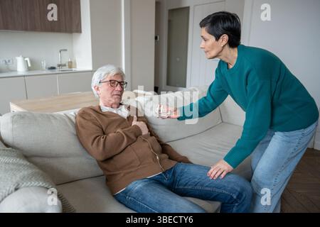 La moglie premurosa aiuta gli anziani che soffrono marito a stringere il petto seduto sul divano, offrendo un bicchiere d'acqua Foto Stock