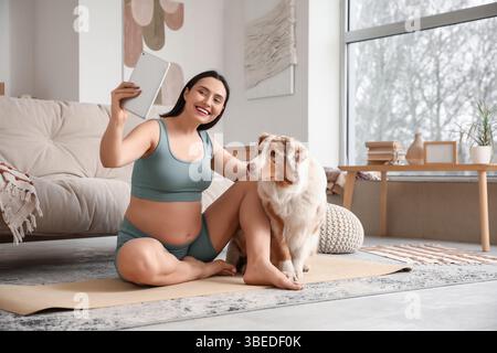 Giovane donna incinta, felice e sportiva, con tablet e simpatico cane pastore australiano che si fa selfie a casa Foto Stock