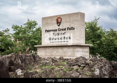 Paterson, New Jersey - Stati Uniti - 24 maggio 2025 cartello di pietra per il Paterson Great Falls National Historical Park si erge tra rocce aspre e vegetazione sotto una nuvolosa sk Foto Stock