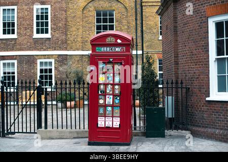 Una classica cabina telefonica rossa coperta di adesivi è visibile in una strada di Londra sullo sfondo di un edificio in mattoni. Foto Stock