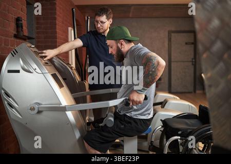 Giovane che assiste un uomo disabile in palestra con attrezzature per esercizi con particolare attenzione all'allenamento di forza e alla riabilitazione. Sedia a rotelle visibile sullo schienale Foto Stock