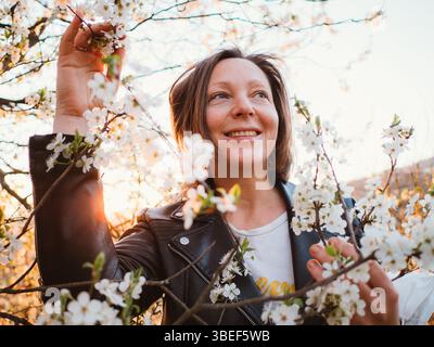 Donna sorridente circondata da ciliegi in fiore all'alba, godendosi l'atmosfera tranquilla. Indossa una giacca di pelle, toccando delicatamente i rami Foto Stock