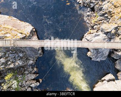Vista aerea con droni: Uno stretto ponte sospeso in legno attraversa un fiume turbolento in un canyon roccioso della natura selvaggia della Norvegia. Foto Stock
