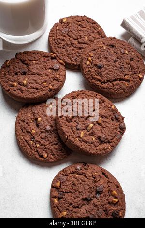 Biscotti con scaglie di cioccolato e un bicchiere di latte su sfondo bianco. Biscotti al cioccolato fondente fatti in casa con nocciole. Foto Stock