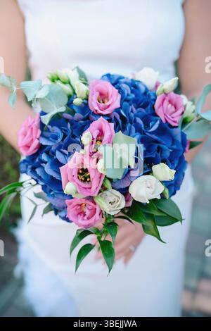 La sposa regge un bouquet elegante caratterizzato da magenta lisianthus, ortensie blu profondo, rose bianche e accentuate da foglie di eucalipto e vegetazione lussureggiante Foto Stock