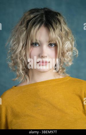Un ritratto accattivante di una giovane donna con capelli biondi ricci, che indossa un maglione giallo senape. Il morbido sfondo blu completa il suo sereno espr Foto Stock