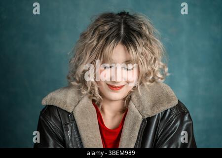 Un ritratto sereno di una giovane donna con capelli ricci e occhi chiusi, sorridente dolcemente. Indossano una giacca in pelle e una camicia rossa, su una b testurizzata Foto Stock