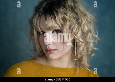 Un ritratto ravvicinato presenta una giovane donna bionda con capelli ricci e bangs, che indossa un maglione giallo. Guarda delicatamente la telecamera, contro un mu Foto Stock