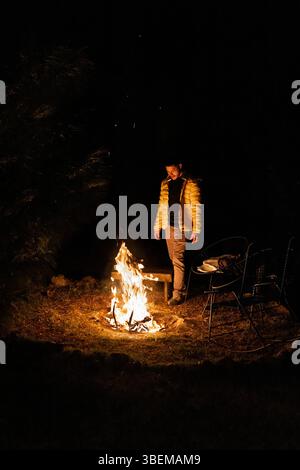 Un uomo sta accanto a un fuoco scoppiettante sotto il cielo notturno in una foresta remota. Il calore del fuoco, circondato dalla natura, crea un sereno e pacifico Foto Stock