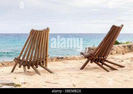 Due sedie a sdraio in legno posizionate invitantemente su una spiaggia sabbiosa a Cozumel, di fronte al mare cristallino dei Caraibi sotto un cielo parzialmente nuvoloso. Foto Stock