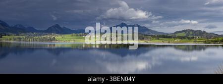 All'alba sul lago Hopfensee, ammira la riva occidentale illuminata dal sole con fattorie, dietro le quali si ritirano le nuvole di tufo, il lago Hopfensee, Ostallgaeu, Allgaeu Foto Stock