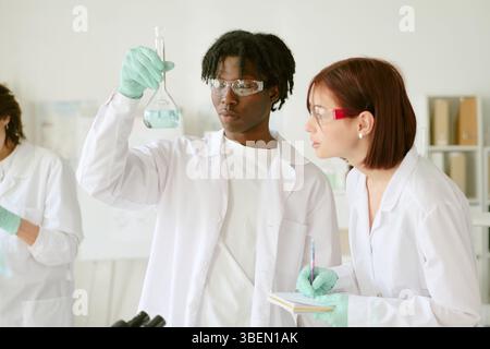 Scienziati impegnati in un laboratorio che esegue esperimenti scientifici. Studente afroamericano che tiene in mano la fiasca mentre donna caucasica osserva con note Foto Stock