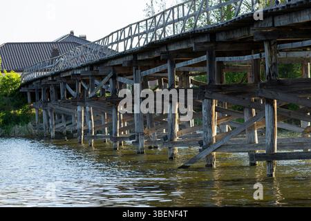 Un affascinante ponte pedonale in legno si estende attraverso il fiume, le sue tavole intemprate forniscono un percorso rustico attraverso la natura. Il flusso sereno del ri Foto Stock