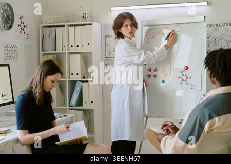 Scienziato che spiega i concetti scientifici sulla lavagna bianca mentre i membri del team prendono appunti in laboratorio. Ricercare i materiali e i diagrammi che circondano Foto Stock