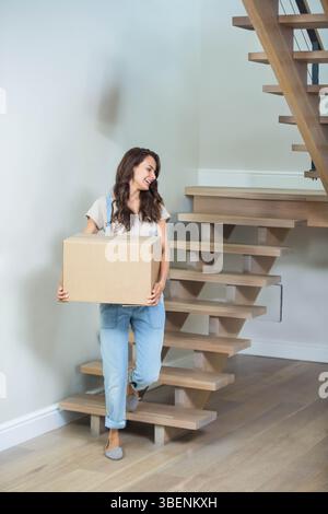 Donna adulta che porta una scatola mobile di cartone mentre sale la scala di legno nel vano scale di casa Foto Stock