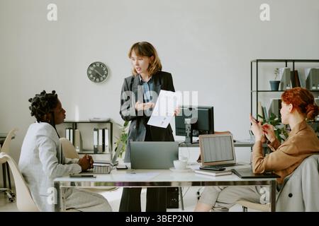 Tre donne che partecipano a riunioni di lavoro intorno a un tavolo con laptop, documenti grafici e orologio sulla parete in background. Parlano e uno è holdi Foto Stock