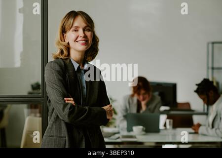 Ritratto di una donna d'affari sorridente in un ambiente d'ufficio moderno con colleghi che lavorano in background, trasudando professionisti e ap Foto Stock