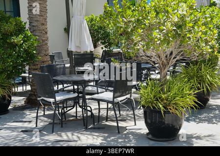 Accogliente area all'aperto con posti a sedere di un ristorante vuoto o di un bistrot circondato da piante su vasi di fiori. Tavoli e sedie neri con cuscini sul patio o sui caffè. Foto Stock