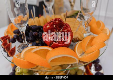 Elegante vassoio di frutta con arance, melograni, uva, fichi e bicchieri da vino su un tavolo bianco, creando un ambiente festoso e lussuoso Foto Stock