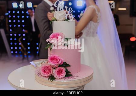 Una torta nuziale rosa a due piani splendidamente decorata, adornata con rose bianche e una torta "MRS", è il punto focale di un gioioso ricevimento di nozze Foto Stock