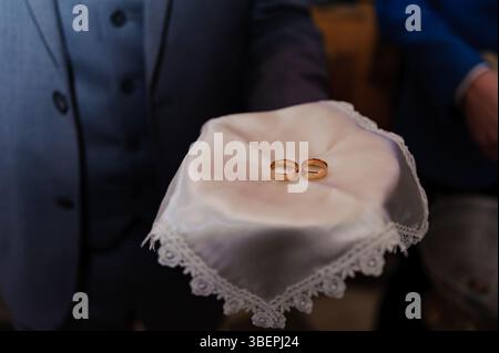 Uno scatto ravvicinato che cattura le fedi nuziali dorate presentate su un delicato fazzoletto in pizzo bianco tenuto da un uomo con una tuta blu, a simboleggiare l'impegno e l'impegno Foto Stock