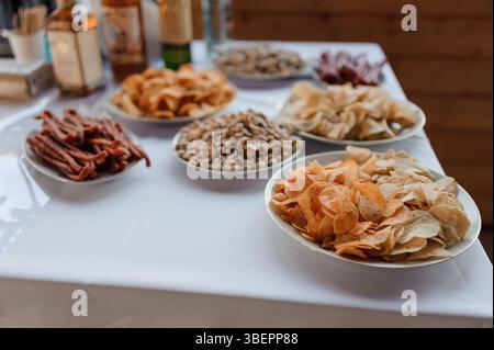 Un vivace assortimento di spuntini salati, tra cui colorato jerkey, varie noci e patatine assortite, esposti su tovaglia bianca durante un evento all'aperto. Foto Stock