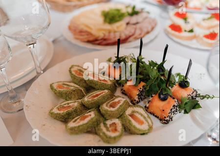 Elegante ambiente da tavola con una varietà di antipasti, tra cui sushi roll e piatti di carne, disposti su una tovaglia bianca, con bicchieri da vino e Foto Stock