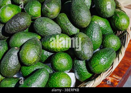 Primo piano di avocado verdi freschi accatastati in un cesto intrecciato presso un mercato locale all'aperto. L'illuminazione naturale ne mette in risalto la consistenza e la maturità Foto Stock