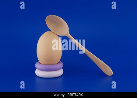 Una disposizione artistica minimalista con un uovo e un cucchiaio di legno su uno sfondo blu vivace. Perfetto per rappresentare la semplicità, la cottura, o. Foto Stock