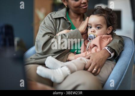 Mamma afroamericana che coccola il bambino con il succhietto seduto sul divano. Scena calda che mostra aderenza e comfort mentre la mamma tiene delicatamente il bambino vicino Foto Stock