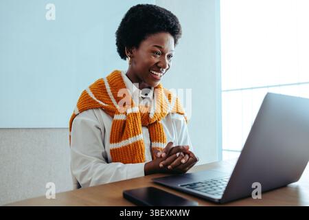 Una donna d'affari nera professionista è impegnata in una riunione online, sorridendo calorosamente mentre interagisce tramite il suo laptop. Sta indossando un maglione arancione A. Foto Stock
