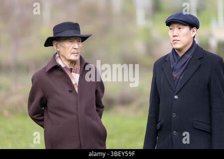 Due uomini asiatici anziani che indossano cappotti e cappelli in piedi nel parco Foto Stock