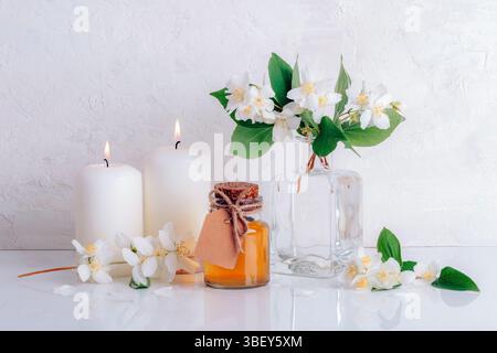Vaso in vetro con olio essenziale di gelsomino, candele e fiori su sfondo bianco. Aromaterapia e trattamento di bellezza. Primo piano. Foto Stock