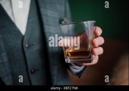 Un uomo ben vestito, probabilmente sulla trentina, vestito con una tuta a quadri grigia testurizzata e camicia bianca, regge con cura un vetro di roccia pieno di ambra scuro Foto Stock