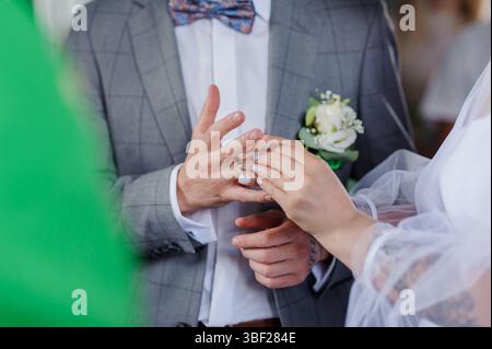 Primo piano di uno sposo con una tuta a scacchi grigia e un cravatta blu, mettendo con cura una fascia nuziale dorata sul dito della sposa durante la cerimonia di nozze Foto Stock